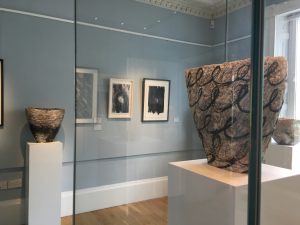 Exhibition of ceramic vessels on white plinths and framed drawings on blue walls