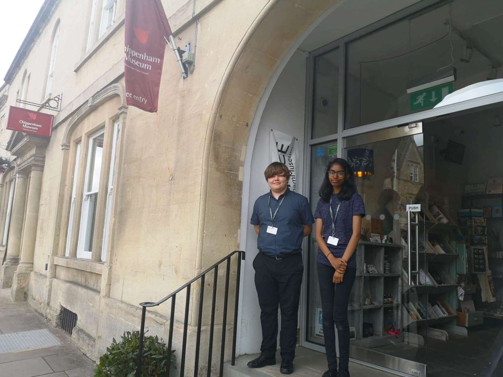 Two people standing on the steps outside Chippenham Museum. A Red flag is on the side of the building, glass doors within an arch way and sandy colour walls.