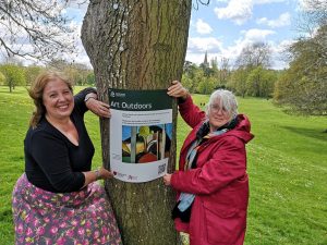 two women stood next to a tree holding an art outdoors banner in a park