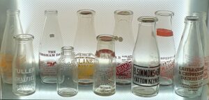 Ten glass milk bottles in a display case, some with cardboard stoppers