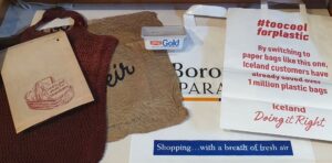 A display case with a selection of paper and plastic shopping bags, a plastic butter tub and a fabric net shopping bag