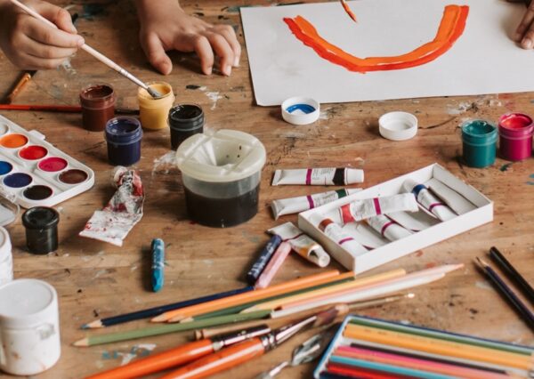 Table top with a range of paints, colouring pencils and pastels, with children's hands doing artwork