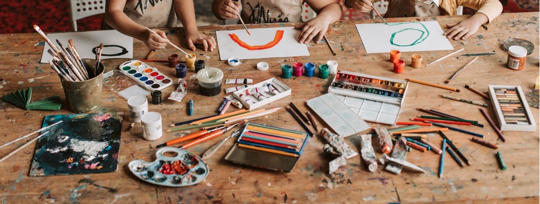 Table top with a range of paints, colouring pencils and pastels, with three children's hands doing artwork
