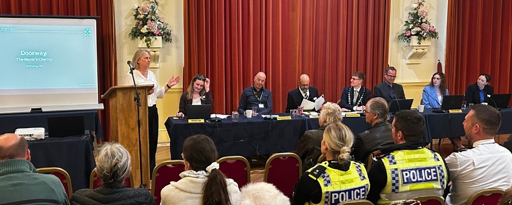 Annual Town Meeting Header Image (10) Photograph of a meeting being held in the town hall with a table set up at the front, a lady talking into a microphone and an audience sat watching