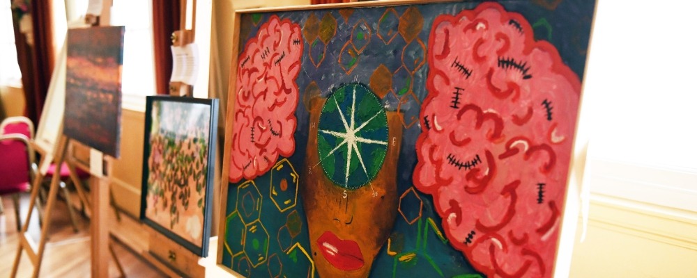 Photograph of a painting which incudes the bottom half of a face, a compass and 2 brains either side with colours such as brown, blue, green, red