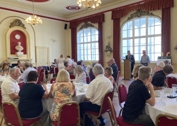 Twinning Association Visit July 2024 Photograph of people sat in the town hall at tables whilst 2 people are stood at the front giving a speech
