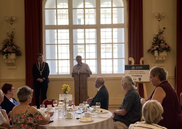 Twinning Association Visit July 2024 Photograph of people in the town hall sat around tables whilst somebody is giving a speech at the front with the mayor