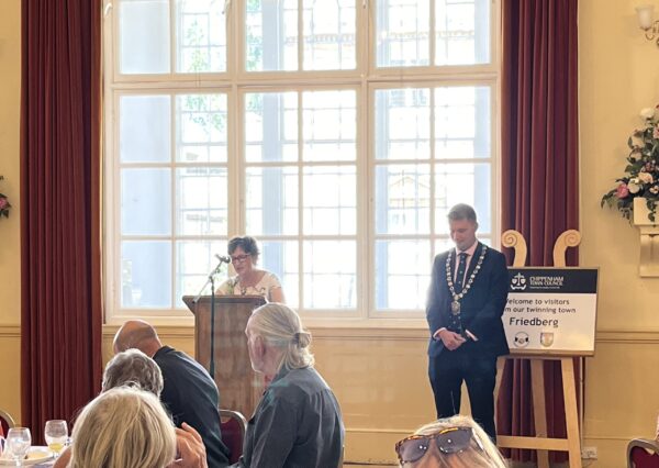Twinning Association Visit July 2024 Photograph of somebody speaking into a microphone with the mayor stood next to them whilst having an audience