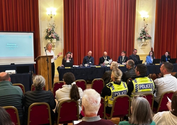 Photograph of people having a meeting in the town hall with someone giving a speech, other people sat at a table and people watching in the audience