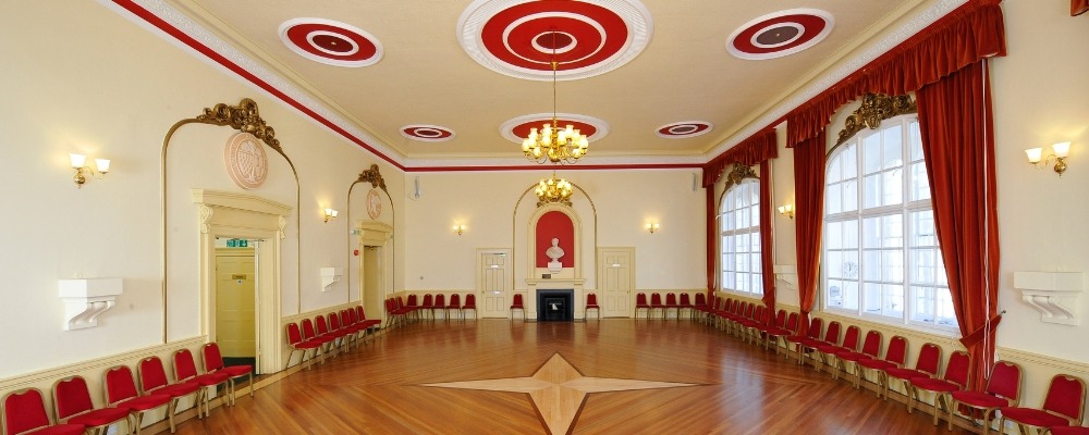 Town Hall Header Image Photograph of the inside of the town hall with red chairs and curtains and hanging lights from the ceiling
