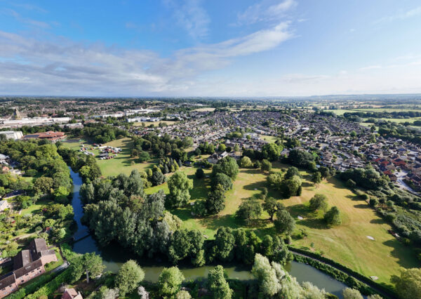 Photograph from above of green fields and trees with a blue sky and lots of houses in the distance