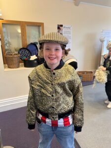 A smiling child wearing a vintage-style cap and gold jacket in a dress-up area.