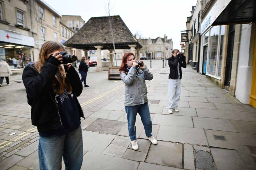 Three young people in Chippenham High taking photographs with the Buttercross in the background.