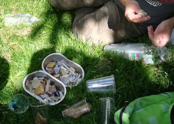 A collection of litter and small found objects laid out on grass beside several plastic cups, with a person sorting through the items outdoors.