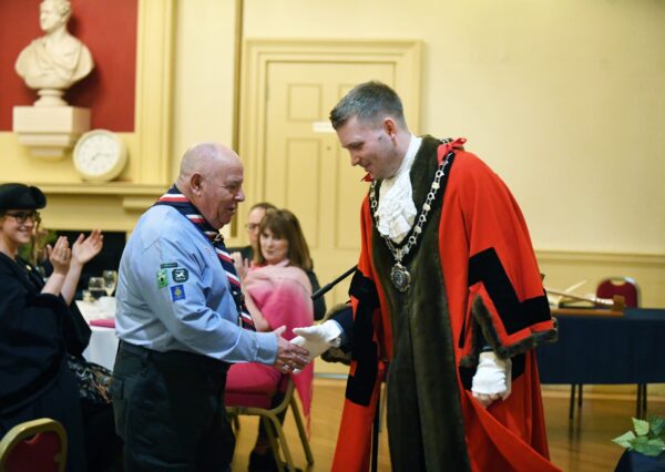 The Mayor Conor Melvin in ceremonial robes smiling and shaking hands with Rodney Jefferies MBE in Chippenham Air Scouts uniform with three other people in the back ground smiling and clapping.