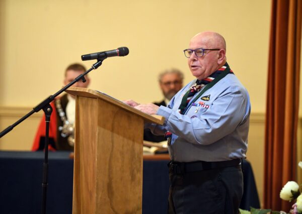 A person in uniform stands at a lectern speaking to the room, two other people in the background behind a blue clothed table.
