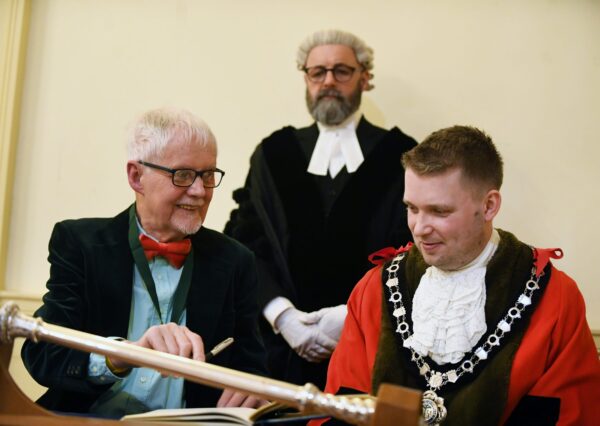 Three people pose for a formal photo: two seated with one in ceremonial robes ready to sign a book. A third person wearing robes of office stands behind and observes.
