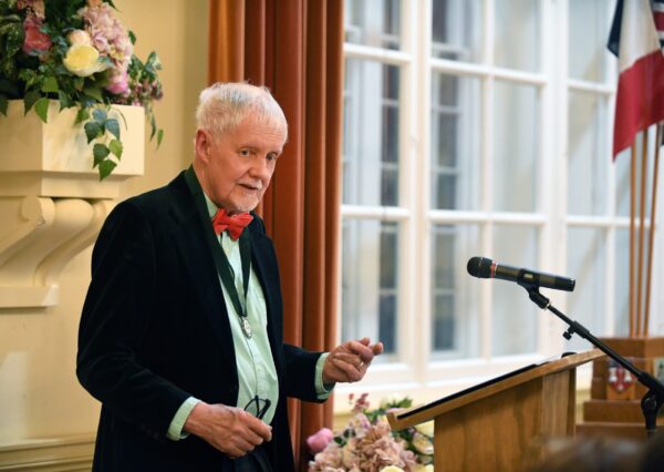 A person stands at a lectern speaking to the room, with a bouquet of flowers and flags in the background.