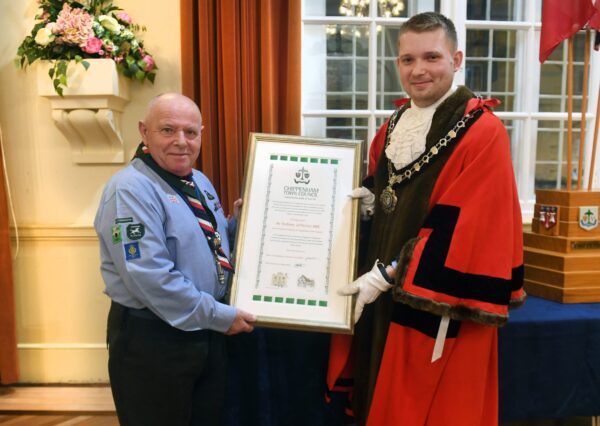 Two standing people smiling one in ceremonial robes and civic chain the other in the Chippenham air scouts uniform both holding a framed award. A bouquet of flowers and flags are in the background.