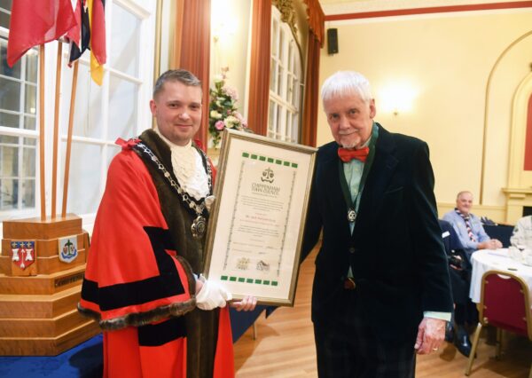 Two people smiling one in ceremonial robes and civic chain both holding a framed award. Two people in the back ground are seated at a table and look on.