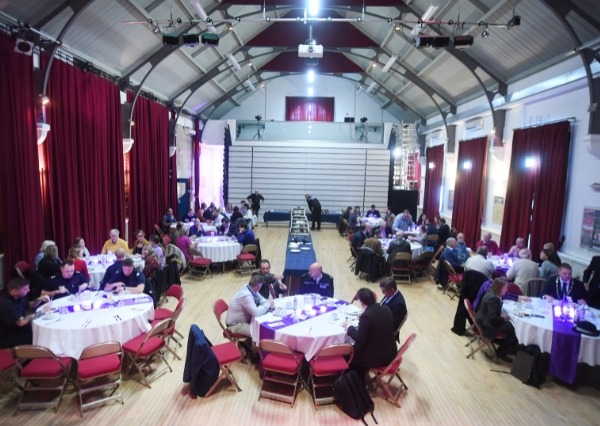 Purple Flag Breakfast A busy Neeld hall with round tables, people gathered in small groups, red stage curtains, and tiered seating at the far end. The space is bright and open .