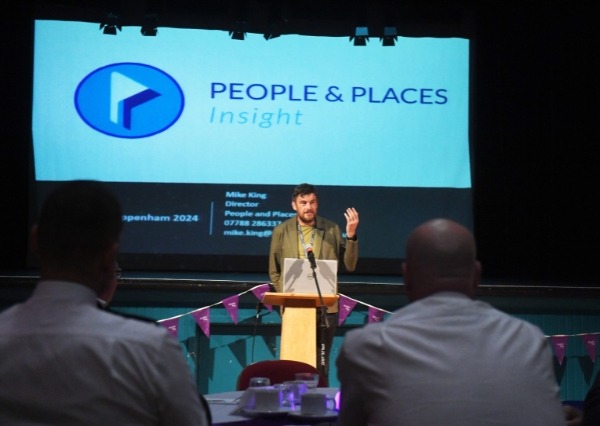 Purple Flag Breakfast A person stands at a lectern giving a presentation, with a large screen behind them displaying the People & Places Insight logo and event details. Audience members are seated in the foreground facing the speaker.