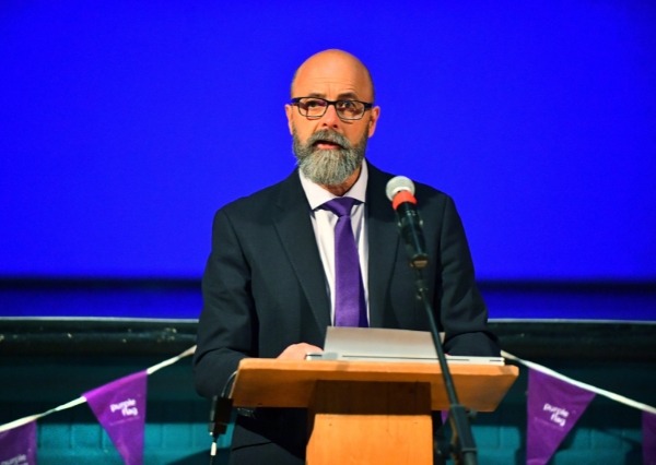 Purple Flag Breakfast A person in a dark suit and purple tie stands at a lectern speaking into a microphone, against a bright blue-lit background with purple event bunting visible below.