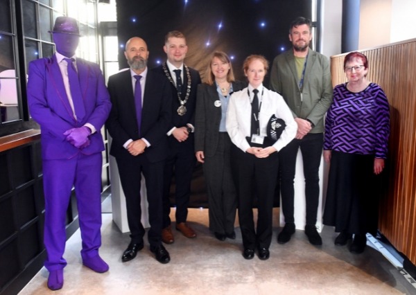 Purple Flag Breakfast A group of people standing together indoors for a posed photo, with a dark backdrop lit by small lights behind them. One person is dressed entirely in purple, while the others are in smart or business-style clothing.