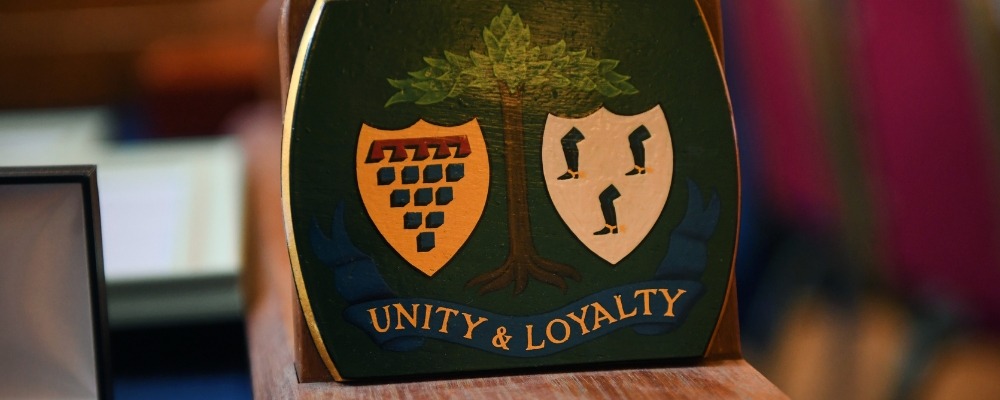 A close-up of a decorative wooden crest featuring two shields, a tree, and the words “Unity & Loyalty” on a blue banner
