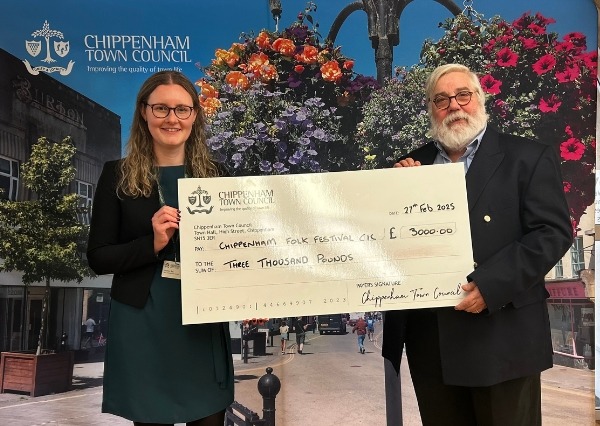 Council Grants Scheme Two people stand smiling in front of a Chippenham Town Council backdrop, holding a large check awarding £3000.00 to Chippenham Folk Festival