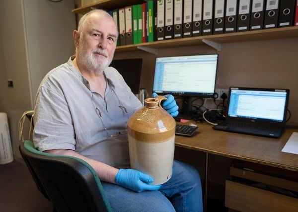 Collections Volunteer A man sitting holding a stoneware brewery jar from the museum collection, with a computer on a desk in the background.