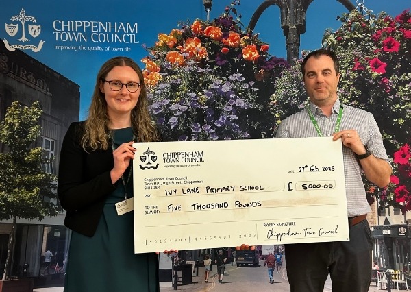 Council Grants Scheme Two people stand smiling in front of a Chippenham Town Council backdrop, holding a large check awarding £5000.00 to Ivy Lane School