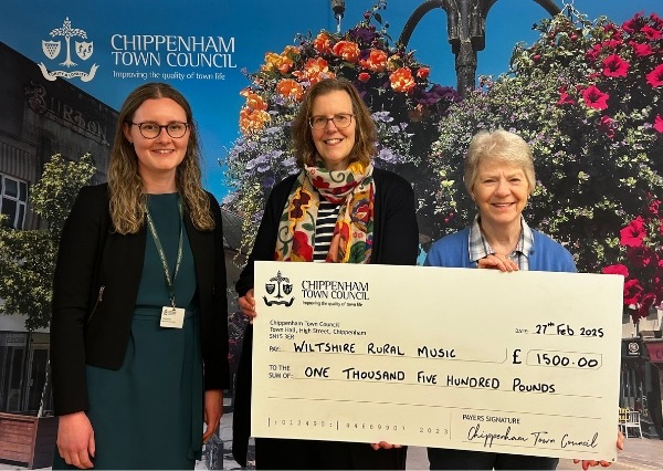 Council Grants Scheme Three people stand smiling in front of a Chippenham Town Council backdrop, holding a large check awarding £1500.00 to Wiltshire Rural Music