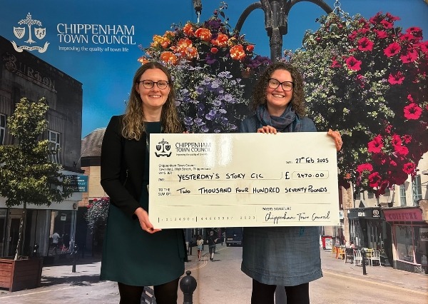 Council Grants Scheme Two people stand smiling in front of a Chippenham Town Council backdrop, holding a large check awarding £2470.00 to Yesterday's Story