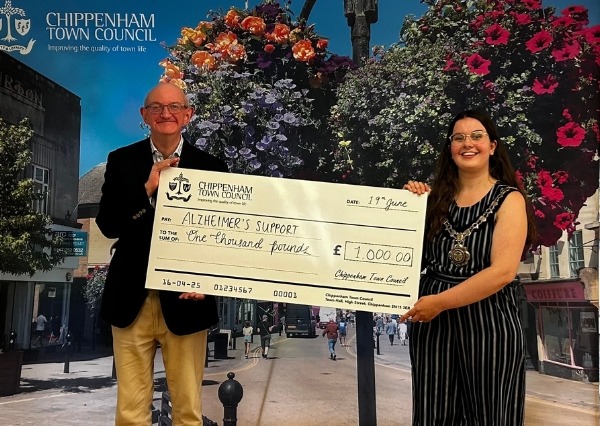 Two people stand smiling in front of a Chippenham Town Council backdrop, holding a large check awarding £1000 to Alzheimers Support Two people stand smiling in front of a Chippenham Town Council backdrop, holding a large check awarding £1000 to Alzheimers Support
