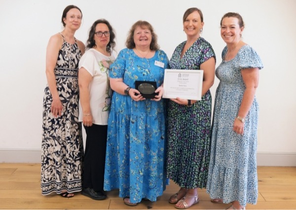 Civic awards Five women stand together indoors, smiling at the camera. Two women in the center hold an award plaque and a framed certificate. They are dressed in colorful dresses and casual clothing.