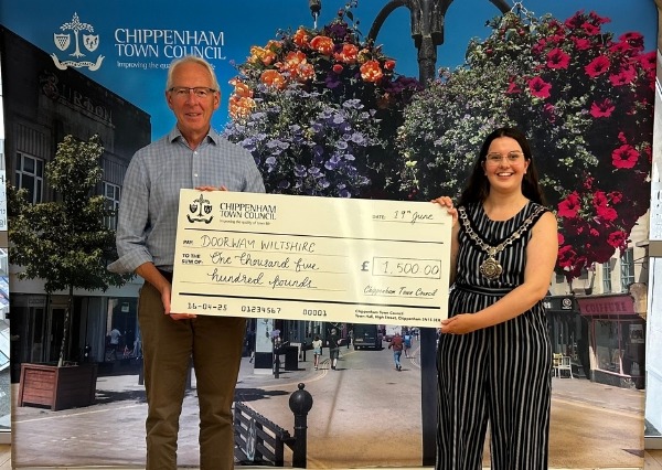 Two people stand smiling in front of a Chippenham Town Council backdrop, holding a large check awarding £1500 to Doorway Wiltshire Two people stand smiling in front of a Chippenham Town Council backdrop, holding a large check awarding £1500 to Doorway Wiltshire