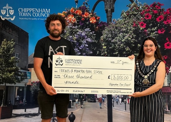 Two people stand smiling in front of a Chippenham Town Council backdrop, holding a large check awarding £3000 to Friends of Monkton Park School Two people stand smiling in front of a Chippenham Town Council backdrop, holding a large check awarding £3000 to Friends of Monkton Park School