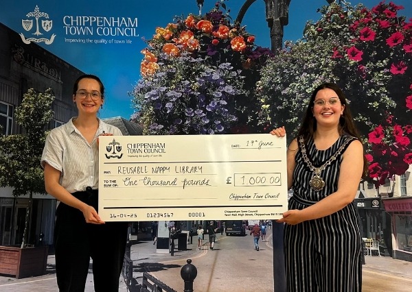 Two people stand smiling in front of a Chippenham Town Council backdrop, holding a large check awarding £1000 Reusable nappy library Two people stand smiling in front of a Chippenham Town Council backdrop, holding a large check awarding £1000 Reusable nappy library