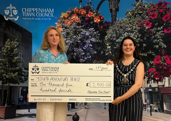 Two people stand smiling in front of a Chippenham Town Council backdrop, holding a large check awarding £1500 to Youth Adventure Trust Two people stand smiling in front of a Chippenham Town Council backdrop, holding a large check awarding £1500 to Youth Adventure Trust