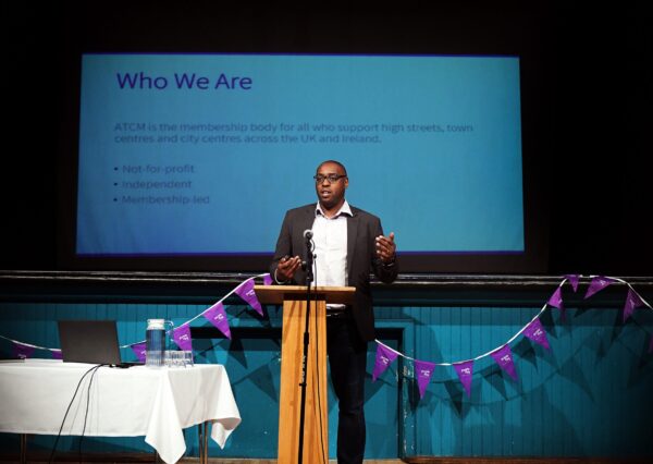 A well dressed man stands at a podium, speaking into a microphone while addressing an audience. A well dressed man stands at a podium, speaking into a microphone while addressing an audience , there is purple flag bunting hanging behind