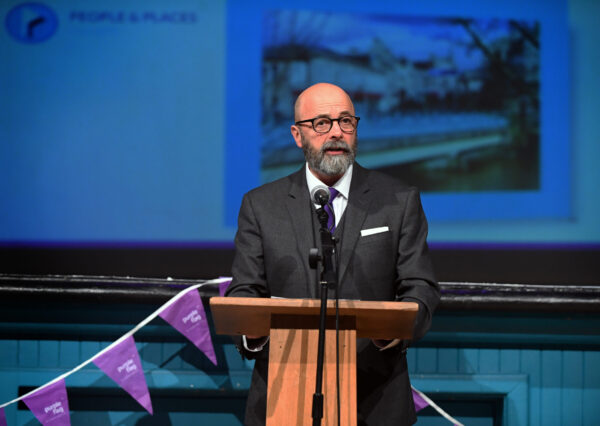 A well dressed man stands at a podium, speaking into a microphone while addressing an audience. A well dressed man stands at a podium, speaking into a microphone while addressing an audience.