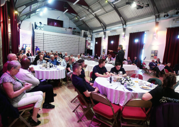 Venue hall full of people sat around tables looking engaged while listening to a talk Venue hall full of people sat around tables looking engaged while listening to a talk