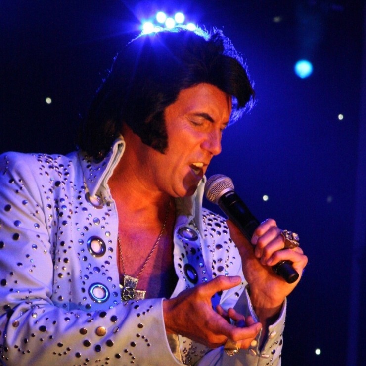 Someone dressed up in a white Elvis suit with black quiffed hair and large side burns singing into a microphone