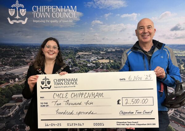 Council Grants Scheme Two people stand smiling in front of a Chippenham Town Council backdrop, holding a large check awarding £2500 to Cycle Chippenham .