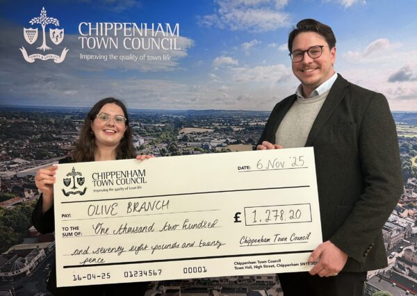 Council Grants Scheme Two people stand smiling in front of a Chippenham Town Council backdrop, holding a large check awarding £1278.20 to Olive Branch.