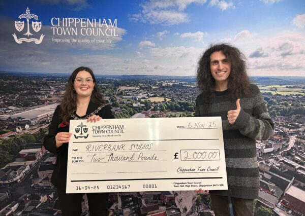 Council Grants Scheme Two people stand smiling in front of a Chippenham Town Council backdrop, holding a large check awarding £2000 to Riverbank Studios.