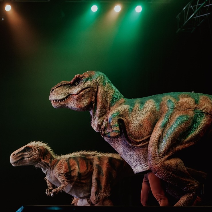 2 humans in costumes T-Rex dinosaurs with orange and green background lighting