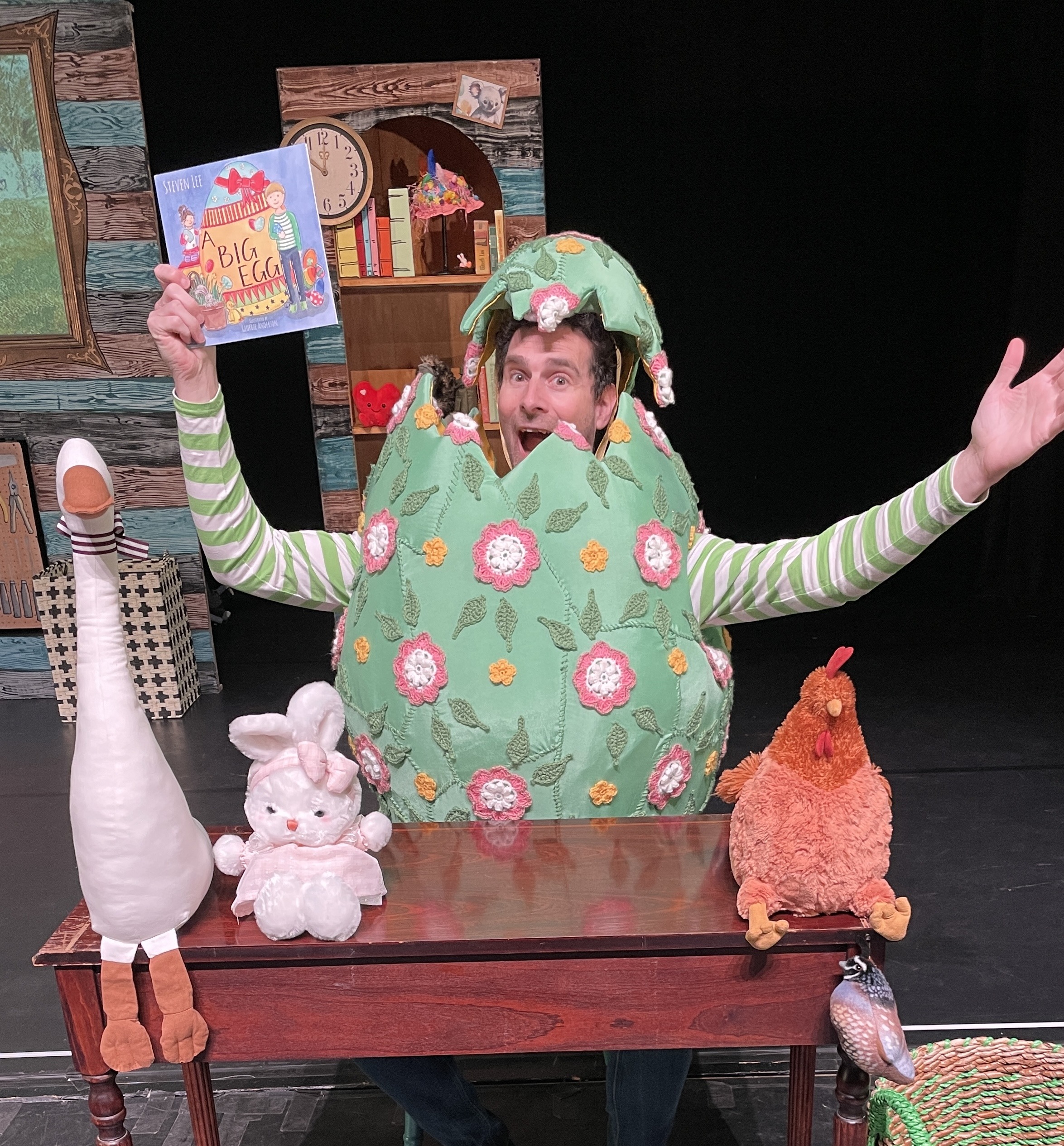 A person on a stage is dressed as a giant cracked green egg, smiling with their arms spread wide while holding up a children's book called "A Big Egg." In front of them are stuffed animals—a goose, a bunny, and a chicken—on a wooden table. The stage is decorated like a cozy room, creating a fun, playful setting for a children's story.