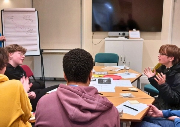 A group of teenage students around a table in conversation with paperwork laid out on tables